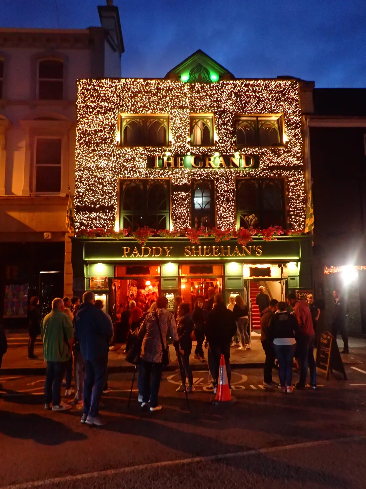 People gather outside Paddy Sheehan’s at The Grand in Killarney, with warm pub lights beneath a blue evening sky.